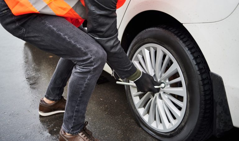 Driver performing a flat tire change by loosening lug nuts with a wrench on a roadside.