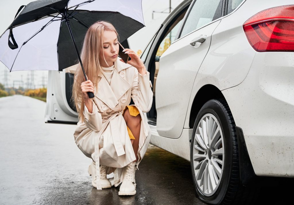 Woman calling for flat tire change assistance beside her car on a rainy day.